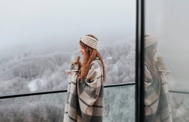 Kareli battaniyeye sarılı mutlu kadın balkon terasında duruyor, sisli kış gününde kahve içiyor. Örülmüş saç bandı takan kadın, bir fincan sıcak içecekle dondurucu dağ sabahının tadını çıkarıyor..