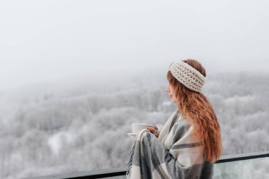 Karla kaplı dağların dingin güzelliğinin tadını çıkarmak için, rahat bir battaniyeye sarılmış kadın sıcak içkinin tadını çıkarıyor. Kadın balkon terasında duruyor, sisli kış gününde sabah kahvesini içiyor..