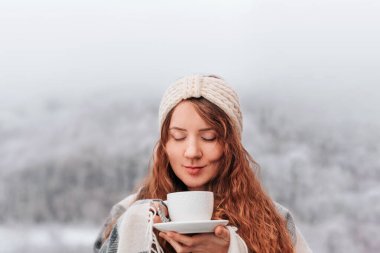 Sıcak, ekose battaniyeli kadın sabah kahvesini içiyor. Karlı kış manzarasının huzurlu huzurunun tadını çıkarıyor. Neşeli kadın sisli kış gününde sıcak içkinin tadını çıkarıyor, hafta sonunun tadını çıkarıyor..