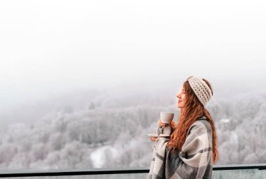 Karla kaplı dağlarla kaplı kış ortamına karşı sıcak içkinin keyfini çıkaran ekose örtülü genç bir kadın. Kadın balkon terasında duruyor, sisli kış gününde sabah kahvesini içiyor..