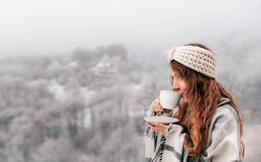 Kareli battaniyeli genç bir kadın, sabah kahvesini içiyor, kış havasına karşı sıcak içkinin tadını çıkarıyor, karla kaplı dağlarla birlikte. Dişi, tatil sezonunda sıcak bir çay yudumluyor..