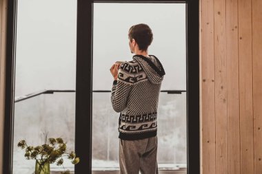 Kereste dağ kulübesinde, pencerenin önünde sabah kahvesi içen sıcak kazaklı bir adam. Erkek, dışarıda karlı manzaraya hayran, doğada tatilin tadını çıkarıyor. Sessiz yaşam tarzı konsepti.