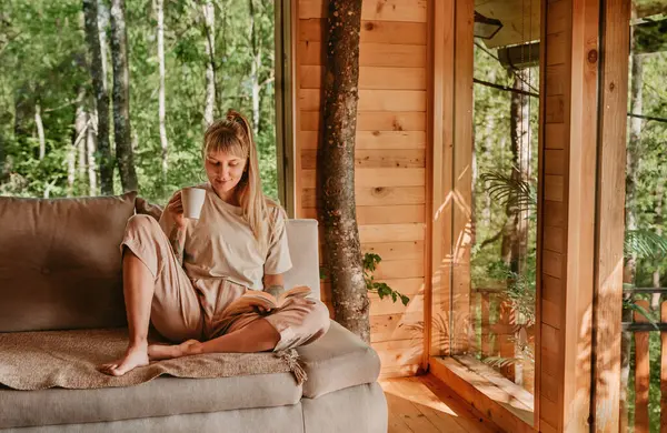 Kanepede oturup kitap okuyan ve orman oduncularının içinde çay ya da kahve içen bir kadın. Haftasonu kaçamağı sırasında sakin ve sakin bir hayatın tadını çıkaran bir kadın, doğadaki ahşap evde dinleniyor..