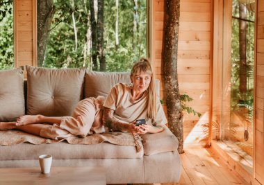 Kadın mesajları okuyor ve kontrol ediyor ya da akıllı telefondaki uygulamayı kullanıyor, odunluktaki kanepede yatıyor. Dişi, rahat bir ağaç evin içindeki kanepede dinleniyor, telefonunu tutuyor, mesaj atıyor ya da internette sörf yapıyor..