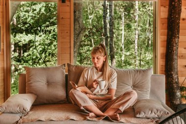 Kanepede oturup kitap okuyan ve orman kulübesinde çay veya kahve içen bir kadın. Kadın, hafta sonu kaçamağı sırasında sakin ve sakin anların tadını çıkarıyor. Doğadaki ahşap evde dinleniyor..