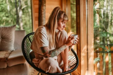 Rahat bir kadın, kabinde rahat bir şekilde oturan, elinde kahve ya da çay fincanı tutan ve pencerenin dışındaki sessiz yeşil ormanı seyreden rahat bir kadın. Kadın dinlenme zamanından zevk alıyor. Doğada yavaş yaşam kavramı.