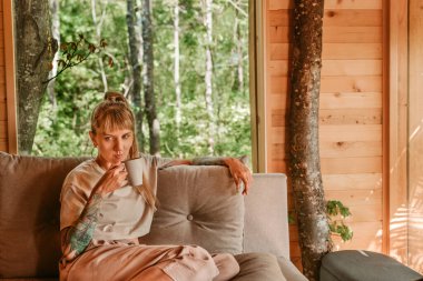 Kanepede oturan bir kadın yeşil ormanla çevrili rahat bir kulübede kahve içiyor. Kadın, hafta sonları ağaç evde eğleniyor. Doğada yavaş düşünceli yaşam kavramı.