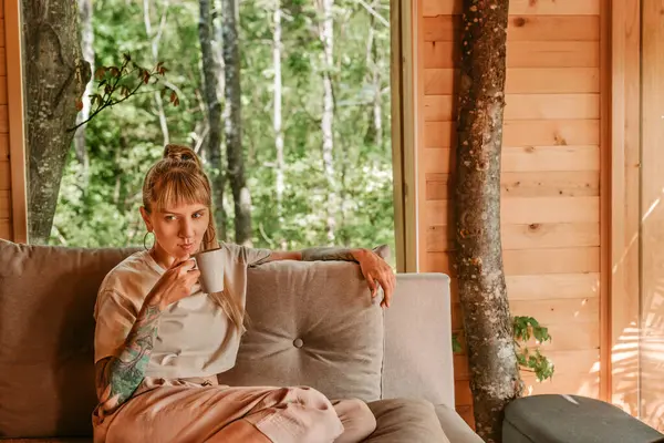 Kanepede oturan bir kadın yeşil ormanla çevrili rahat bir kulübede kahve içiyor. Kadın, hafta sonları ağaç evde eğleniyor. Doğada yavaş düşünceli yaşam kavramı.