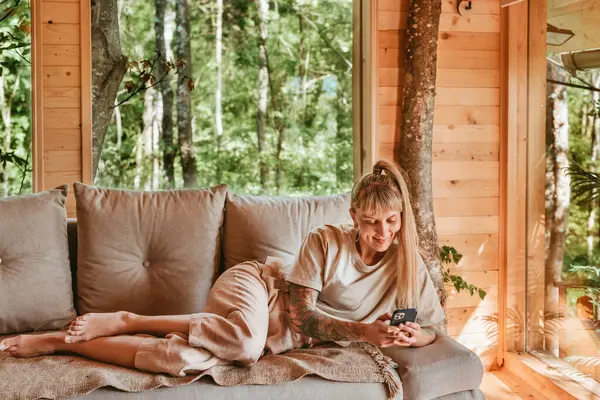 Bir kadın mesajları okuyor ve kontrol ediyor ya da akıllı telefonda uygulama kullanıyor, odunluktaki kanepede dinleniyor. Orman ağaç evinde kanepede uzanmış gülümseyen bir kadın telefonunu tutuyor, mesaj atıyor ya da internette sörf yapıyor..