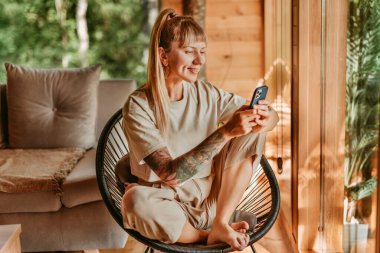 Kadın mesajları okuyor ve kontrol ediyor ya da akıllı telefondaki uygulamayı kullanıyor, kütük kulübesindeki sandalyede dinleniyor. Gülümseyen bir kadın ağaçlık evde rahatça oturuyor telefonla, mesajlaşıyor ya da internette sörf yapıyor..