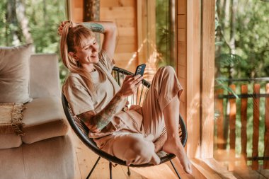 Kadınlar tatil sırasında odunlukta akıllı telefonuyla iletişim kuruyor. Konforlu ağaç evde rahat bir sandalyede dinlenen kadın, mesajları okuyor ve kontrol ediyor, ya da akıllı telefonlarda uygulama kullanıyor.