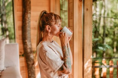 Kadın sabah kahvesinin tadını çıkarıyor, pencerenin önünde dikiliyor. Sıcak, ahşap kulübede içen kadın tişörtü, hafta sonu orman ağaç evinde yapılan kaçamağa hayran. Doğada yavaş yaşam kavramı.