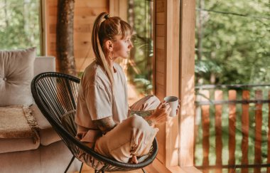 Kadın, kahve içerken doğal manzaranın tadını çıkarıyor, rahat bir kulübede pencere kenarında dinleniyor. Sandalyede oturan bir kadın, hafta sonu orman ağaç evinde kaçamağın tadını çıkarıyor. Doğada yavaş yaşam kavramı.