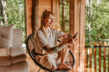 Kız tatil sırasında kulübede akıllı telefonunu kullanarak iletişim kuruyor. Konforlu ağaç evde rahat bir sandalyede oturup mesajları okuyup kontrol eden ya da akıllı telefonunda uygulama kullanan bir kadın..