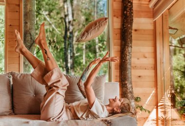 Mutlu bir kadın kanepeye uzanmış, bacakları havada, yastık fırlatıyor, orman kulübesinde eğleniyor. Kadın, hafta sonları ağaç evde eğleniyor. Eğlence ve doğa kavramına kaçmak.