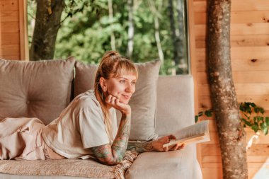 Dağ kulübesinde kitap okumaktan hoşlanan bir kadın, romantik bir ifadeyle kanepede uzanmış, elinde kağıt kitapla. Güzel bir kadın hafta sonu kaçamağı sırasında sakin ve yoğun bir hayatın tadını çıkarıyor..