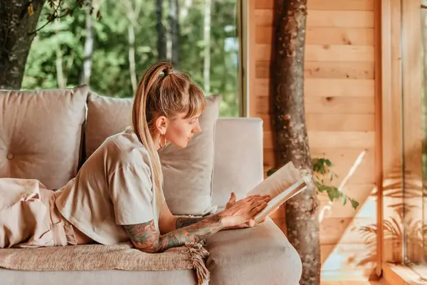 Dağ kulübesinde kitap okumaktan zevk alan bir kadın, heyecan verici romanıyla kanepede uzanıyor. Kadın, orman ağaç evinde tatilde kâğıt kitapla rahatlıyor. Yoğun yaşam konseptinde sakin anlar.