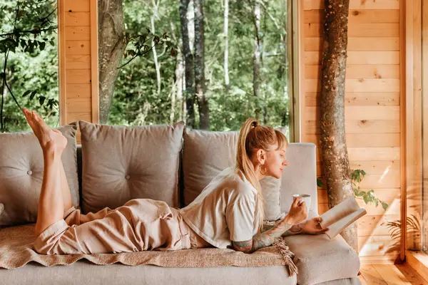 Dağ kulübesinde kitap okumaktan zevk alan bir kadın bir bardak içki ve kağıt kitapla kanepede uzanıyor. Ormandaki ağaç evde tatilde dinlenen bir kadın. Doğada yavaş düşünceli yaşam kavramı.