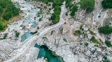 Nehrin üzerindeki eski taş köprüde eşeğe binen kadın. Vlora 'daki Brataj Köprüsü' nün hava aracı görüntüsü, Osmanlı döneminden kalma kültürel anıt. Arnavutluk 'a seyahat, doğa ve kültürü keşfetmek.