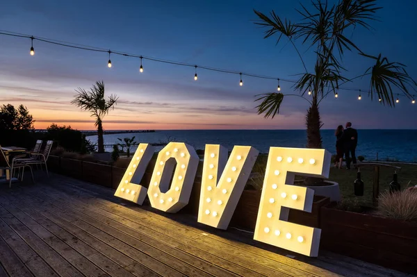 wedding hall interiors and decorations illuminated love inscription against the background of the evening sky with palm trees, a sea bay and a couple in love in the background during the blue hour