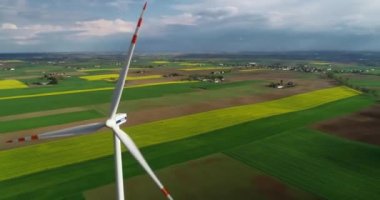 Güneşli bir günde, bahar tarlalarının arka planında çalışan bir rüzgar türbininin ve çiçek açan kanolanın, çarpıcı bir gökyüzünün uzaktan çekimleri. Rüzgar türbini seviyesinde uçuş, yakın plan..