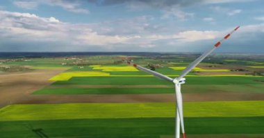 Güneşli bir günde, bahar tarlalarının arka planında çalışan bir rüzgar türbininin ve çiçek açan kanolanın, çarpıcı bir gökyüzünün uzaktan çekimleri. Rüzgar türbini seviyesinde uçuş, yakın plan..