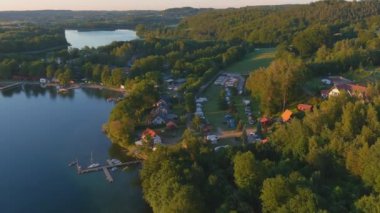 Yaz öğleden sonra Polonya 'daki Kashubian köyü ve göllerinin hava manzarası. Altın saat, kamp, tekneler ve gölde iskele. İleri uçuş çekimi.