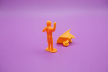 Mor arka planda el arabası oyuncağı ve minyatür işçi izole edilmiş. Sarı el arabası ve oyuncak işçileri ellerini kaldırıyor..