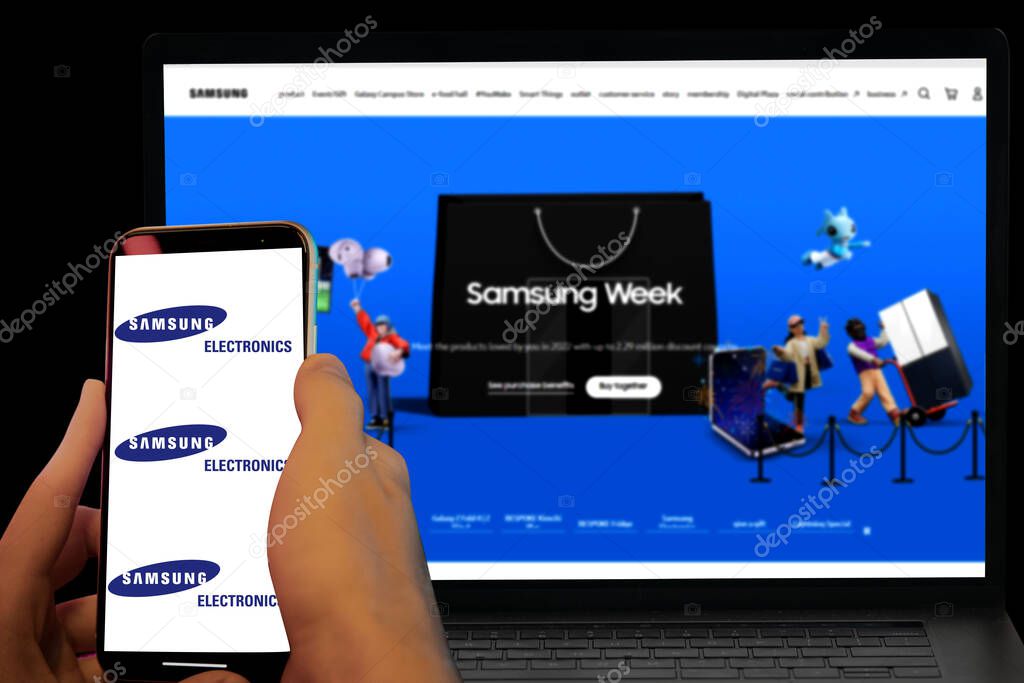 Smartphone displayed Samsung electronics south Korean company logo and their website displayed in blurred laptop screen. illustrative editorial.