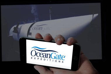 Ocean Gate logo in holding mobile screen and Ocean gate titanic submarine picture in blurred background. clipart