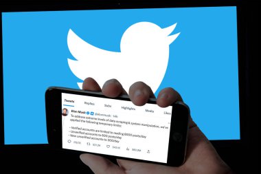 Mobil ekran ve twitter logolarını bulanık dizüstü bilgisayar arka planında tutan Twitter sayfası. Elon Musk Twitter gönderilerini okumak için geçici sınırlar açıkladı.
