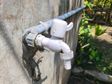 Beyaz plastik bir su musluğu, yıpranmış beton bir duvara tutturulmuş gri bir boruya görünür şekilde bağlıdır. Arka plan, gün ışığında bulanık yeşil yaprakları gösteriyor..