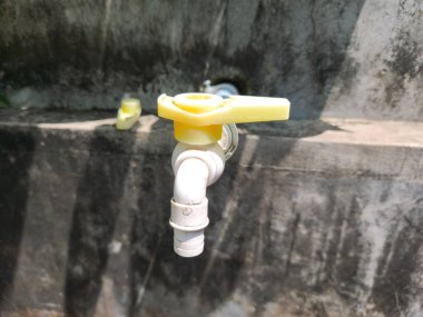 Sarı kolu olan beyaz plastik bir musluk kaba, koyu beton bir duvara monte edilir ve güneş ışığıyla aydınlatılır..