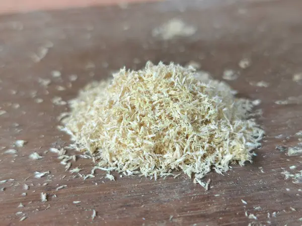 Kahverengi ahşap bir yüzey üzerinde duran açık renkli küçük bir odun talaşının yakın plan çekimi. Küçük parçalar etrafa saçılmış..