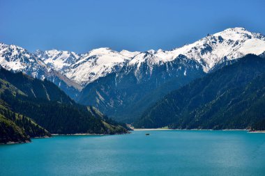 Tianchi Gölü 'nün Tianshan Dağları' ndaki güzel manzarası, Xinjiang, uzakta karla kaplı dağlar, ve önünde büyük ve yeşil Tianhu Gölü, gölde seyahat gemileri, güzel patikalarda oyalanmayı seven turistler.