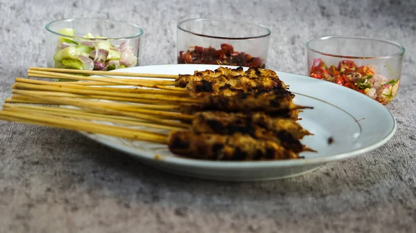 Turşu, soya sosu ve sambal matah ile servis edilen beyaz tabakta satay. Etrafı yiyecek ve içecekle çevrili. Tazeliği aşikar. Göz meleği 