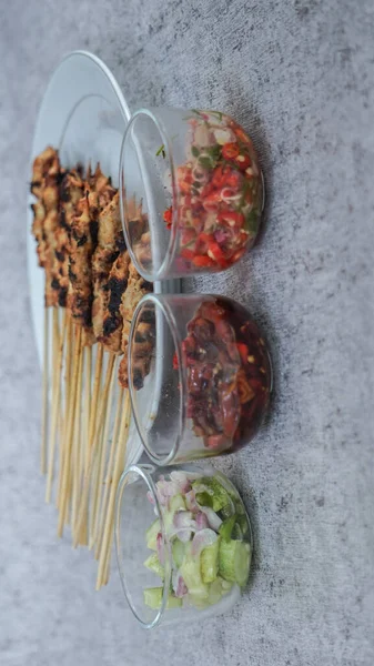 Turşu, soya sosu ve sambal matah ile servis edilen beyaz tabakta satay. Etrafı yiyecek ve içecekle çevrili. Tazeliği aşikar. Potrait Kuşbakışı 