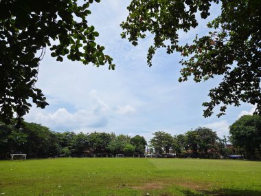 Parçalı bulutlu bir gökyüzünün altında ağaçlarla çevrili yemyeşil bir alan.