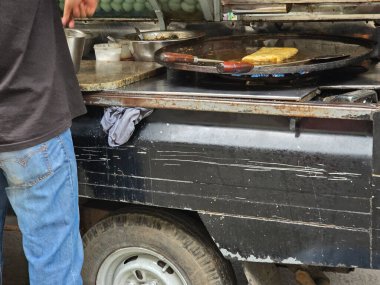 Siyah gömlekli ve kot pantolonlu bir sokak satıcısı açık havada eski siyah yemek arabasında büyük bir tepsi kullanarak kızarmış yumurta martabak hazırlıyor.
