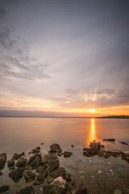 Sabah plajda. Gün doğumu, kayalık bir sahilde çakıl taşı plajı. Gökyüzü bir körfezin suyuna yansıyor. Akdeniz manzarasının doğal manzarası, Vir, Zadar, Dalmaçya, Hırvatistan, Adria