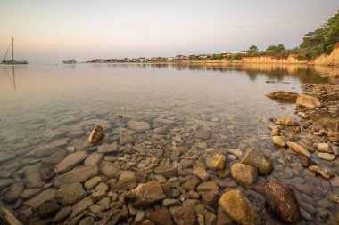 Sabah plajda. Gün doğumu, kayalık bir sahilde çakıl taşı plajı. Gökyüzü bir körfezin suyuna yansıyor. Akdeniz manzarasının doğal manzarası, Vir, Zadar, Dalmaçya, Hırvatistan, Adria