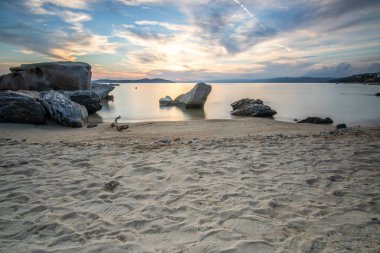 Kumlu bir sahildeki kayaların arasından gün batımına bak. Manzara, Ouranoupoli, Selanik, Orta Makedonya ve Yunanistan kıyılarındaki engin deniz manzarası ile çekildi