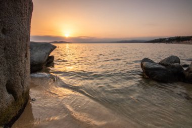 Kumlu bir sahildeki kayaların arasından gün batımına bak. Manzara, Ouranoupoli, Selanik, Orta Makedonya ve Yunanistan kıyılarındaki engin deniz manzarası ile çekildi
