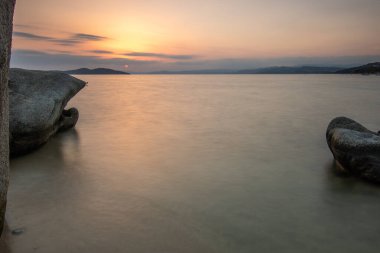 View through rocks on a sandy beach into the sunset. Landscape shot with a view to the horizon over the wide sea on the coast of Ouranoupoli, Thessaloniki, Central Macedonia, Greece clipart