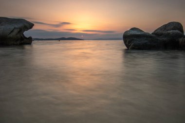 Kumlu bir sahildeki kayaların arasından gün batımına bak. Manzara, Ouranoupoli, Selanik, Orta Makedonya ve Yunanistan kıyılarındaki engin deniz manzarası ile çekildi