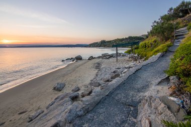 Kumlu bir sahildeki kayaların arasından gün batımına bak. Manzara, Ouranoupoli, Selanik, Orta Makedonya ve Yunanistan kıyılarındaki engin deniz manzarası ile çekildi