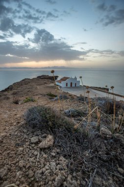 Small white church in the typical architectural style of Greece. She stands on a tree surrounded by the Mediterranean and in the sunrise. Chapel of Agios Nikolaos, Ionian Islands, Greece clipart