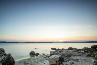 Kumlu bir sahildeki kayaların arasından gün batımına bak. Manzara, Ouranoupoli, Selanik, Orta Makedonya ve Yunanistan kıyılarındaki engin deniz manzarası ile çekildi