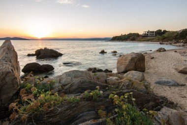 Kumlu bir sahildeki kayaların arasından gün batımına bak. Manzara, Ouranoupoli, Selanik, Orta Makedonya ve Yunanistan kıyılarındaki engin deniz manzarası ile çekildi