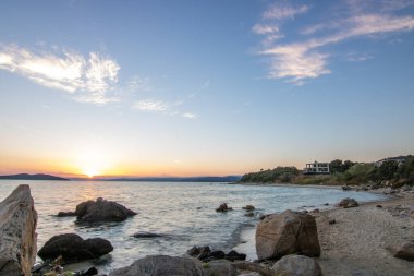 Kumlu bir sahildeki kayaların arasından gün batımına bak. Manzara, Ouranoupoli, Selanik, Orta Makedonya ve Yunanistan kıyılarındaki engin deniz manzarası ile çekildi